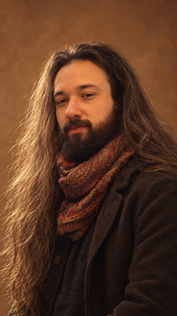 Portrait d’Enzo Brocard, cheveux longs et barbe, portant une écharpe à motifs et une veste sombre, sur un fond brun.